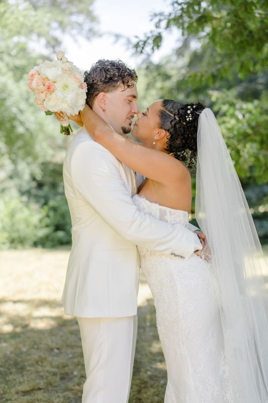 La mariée enlace le marié en costume blanc pour l'embrasser, son long voile flottant