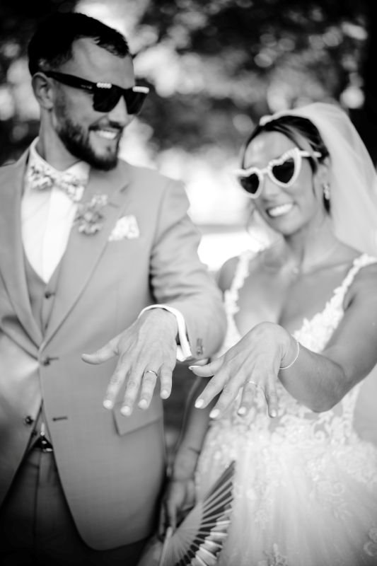 En noir et blanc, les mariés en lunettes de soleil montrent leurs alliances