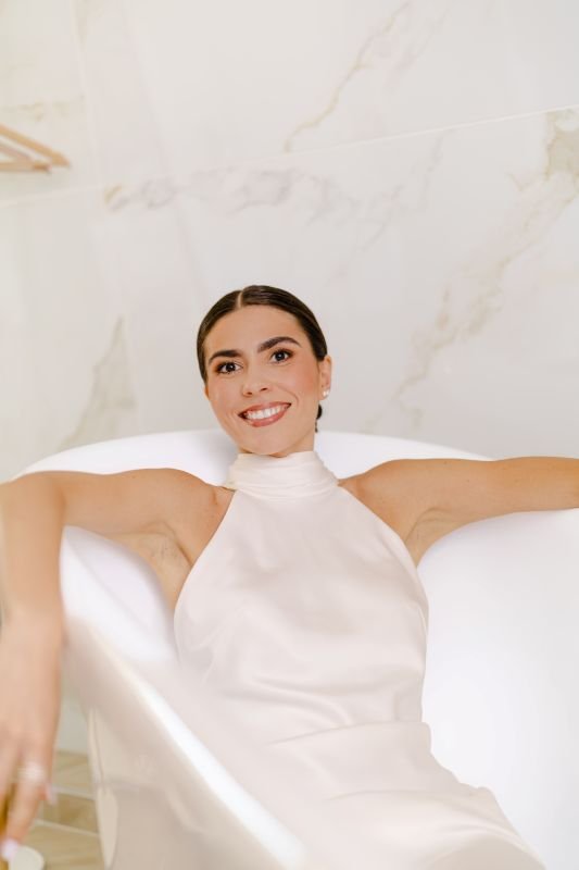 La mariée en robe à col montant, allongée en souriant dans une baignoire en marbre blanc