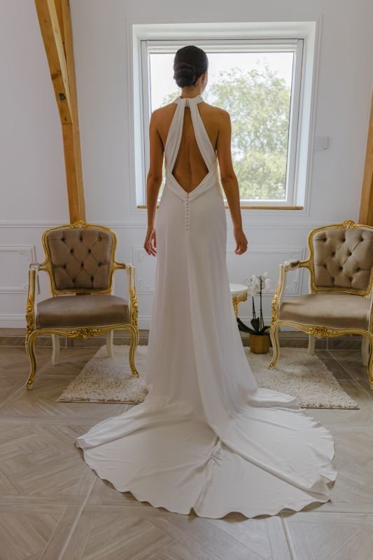 La mariée de dos face à une fenêtre, robe dos nu et longue traîne étalée sur le parquet, entre deux fauteuils dorés