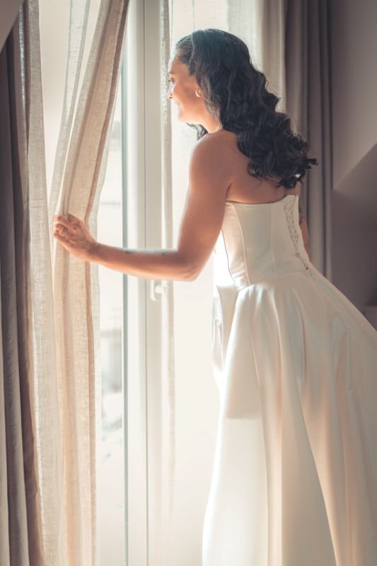 La mariée en robe bustier écarte le rideau d'une fenêtre baignée de lumière