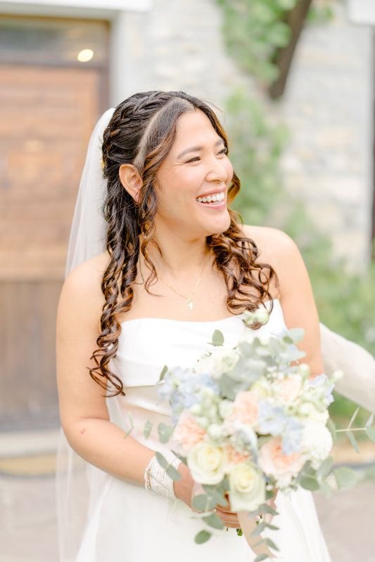 La mariée éclate de rire, bouquet pastel à la main, devant une porte en bois