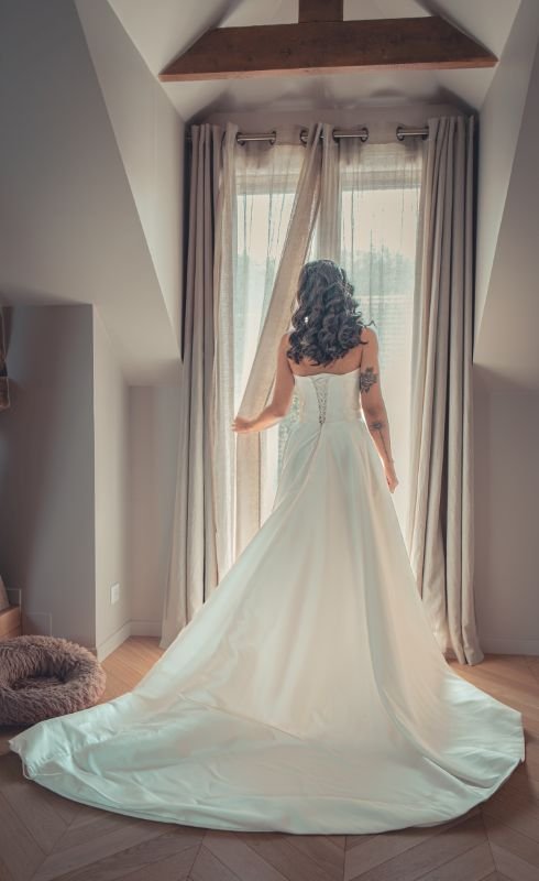 La mariée de dos devant une fenêtre aux rideaux tirés, la traîne de sa robe déployée au sol