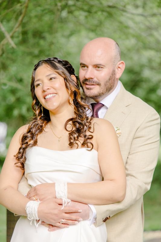 Le marié enlace la mariée par derrière, tous deux souriants dans un jardin