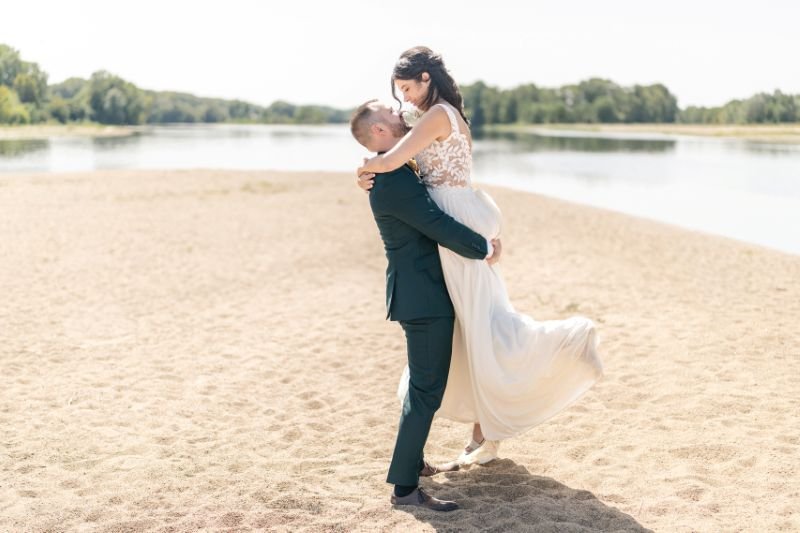 Sur une plage au bord d'un lac, le marié soulève la mariée