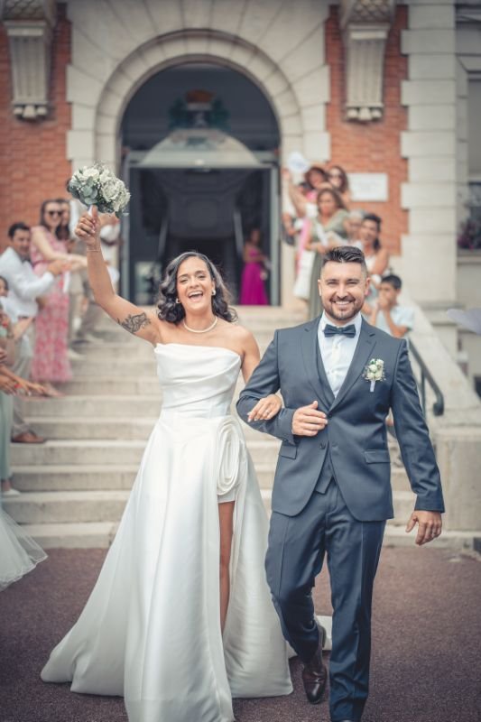 Les mariés descendent un perron, la mariée lève son bouquet sous les applaudissements des invités